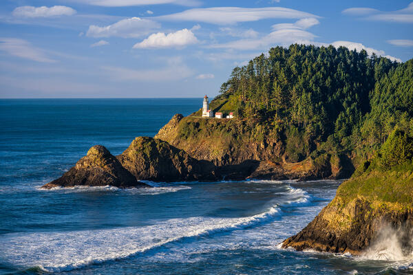 Oregon Coast landscape scene of Heceta Head lighthouse with blue skies and waves coming into shore against the cliffs