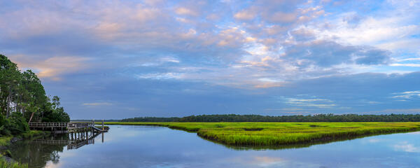 A calm river reflecting a dock and the sky as the river runs through a green marsh in Florida