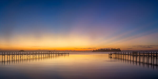 Florida panoramic landscape scene of sunrise sunbeams shooting up from the horizon over two docks and reflecting in the calm water